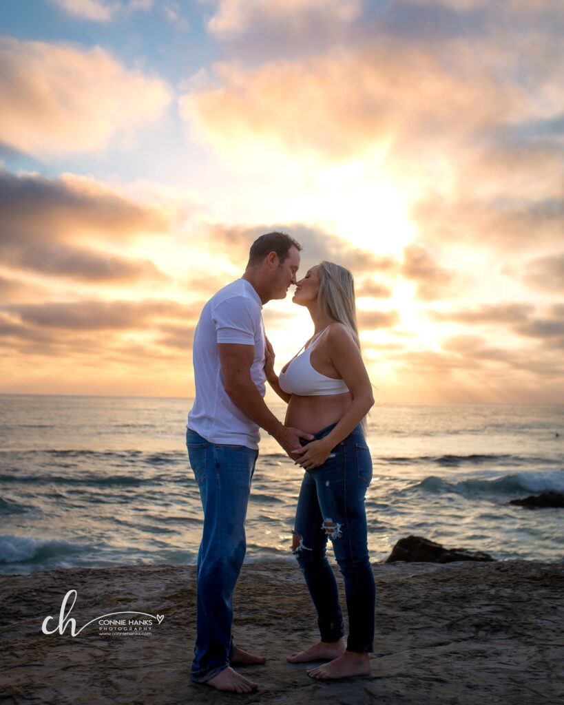 Maternity photos at Windansea Beach, La Jolla. Couple on cliffs along beach with painterly sunset skies.