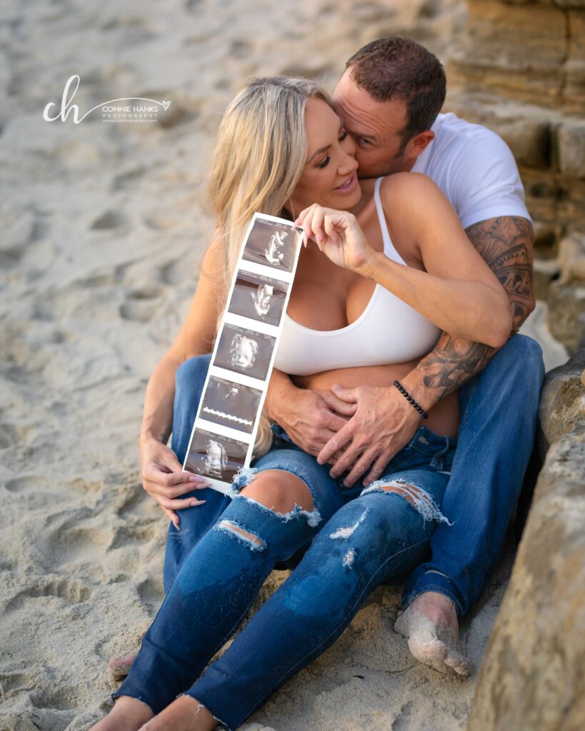 Maternity photos at Windansea Beach, La Jolla. Couple sitting in sand with ultrasound photo strip.