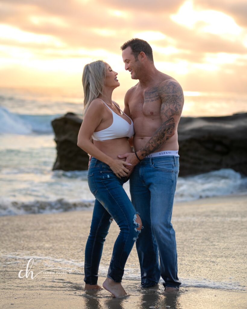 Maternity photos at Windansea Beach, La Jolla. Couple on beach in water with painterly sunset skies.