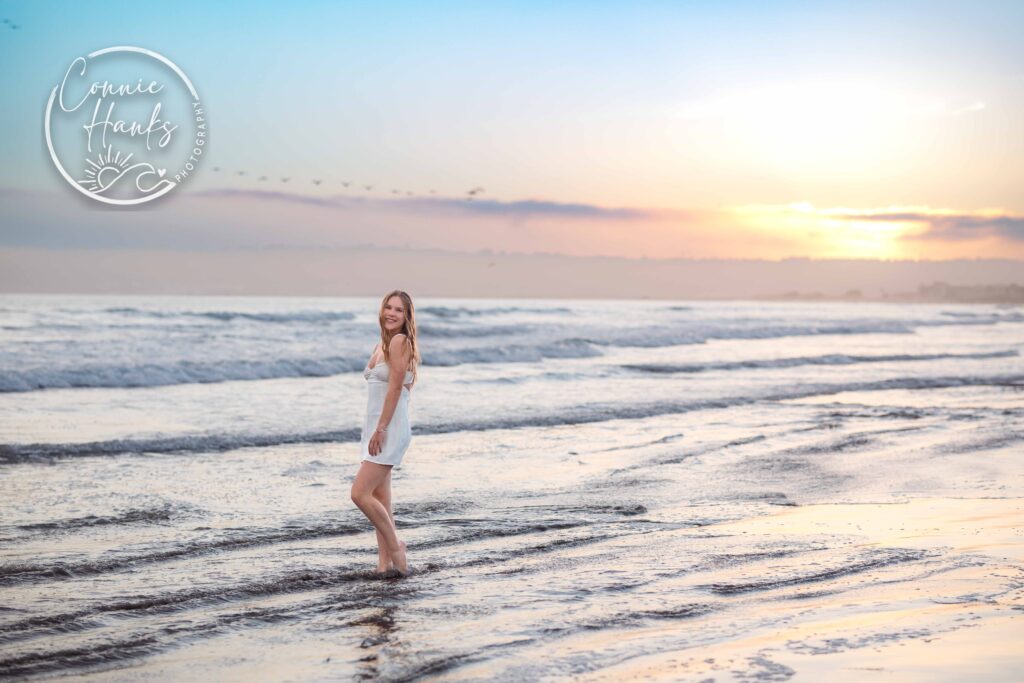 Senior photos at Coronado. Girl in ankle high water at beach with pastel sunset behind her.